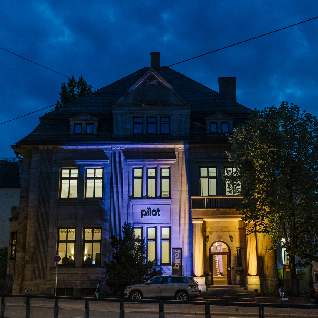 Historisches Gebäude bei Nacht, beleuchtet mit blauem und gelbem Licht, mit dem Schriftzug „pilot“.