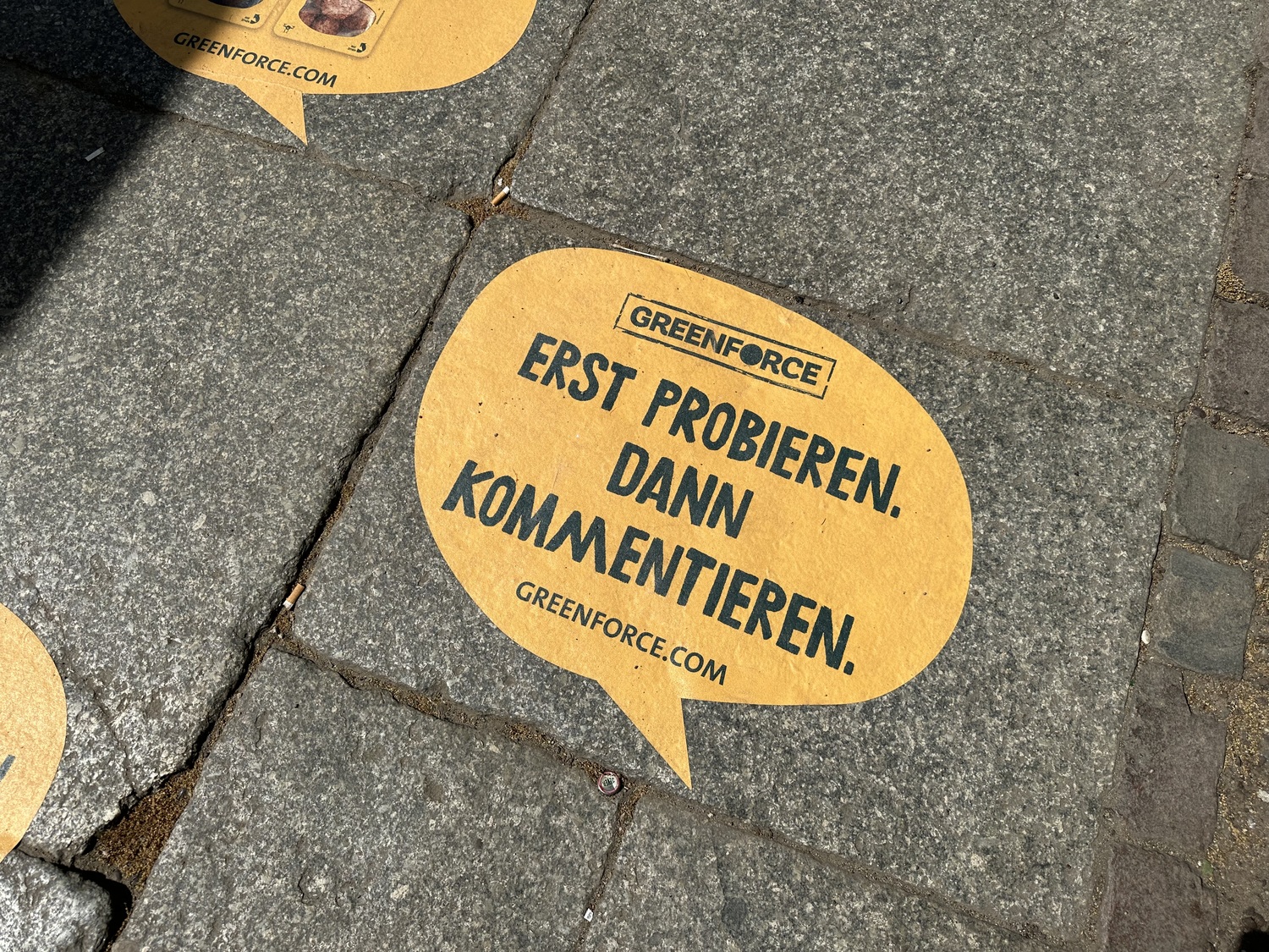 Ein Werbeslogan von Greenforce auf dem Gehweg: "Erst probieren. Dann kommentieren."