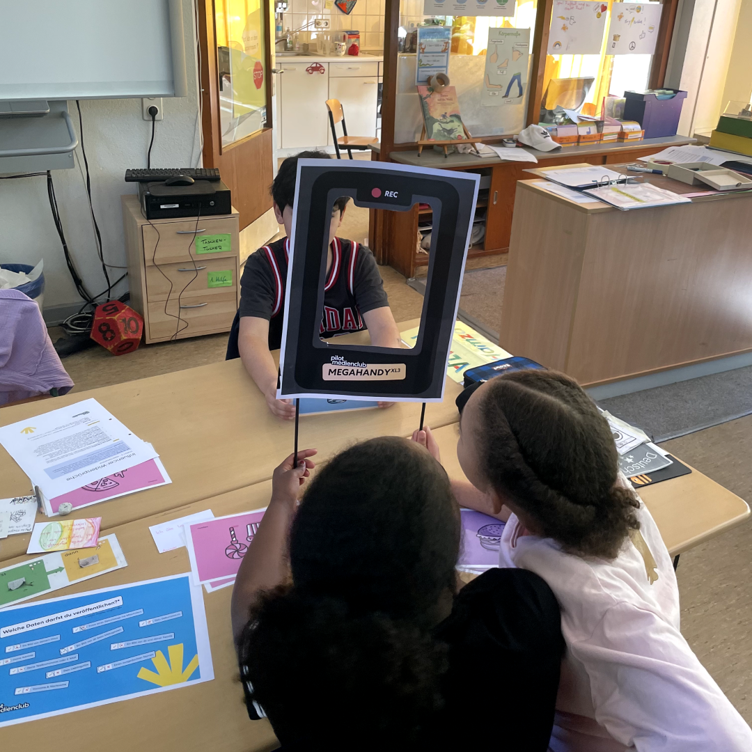 Zwei Kinder halten einen Papprahmen hoch, der wie ein Handybildschirm aussieht, in einem Klassenzimmer.