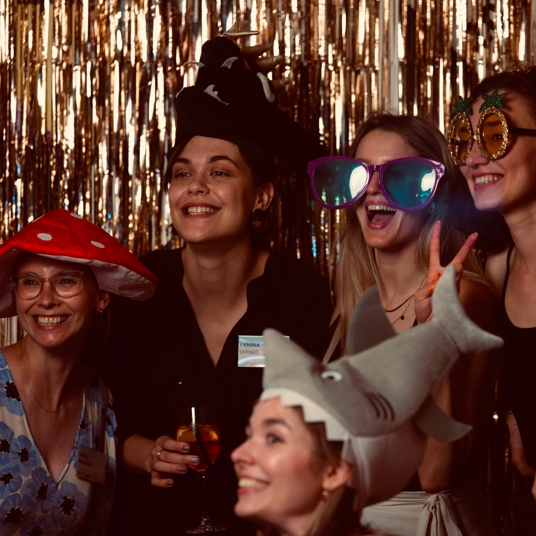 Eine Gruppe von lachenden Frauen trägt lustige Hüte und übergroße Brillen vor einem glitzernden Hintergrund.