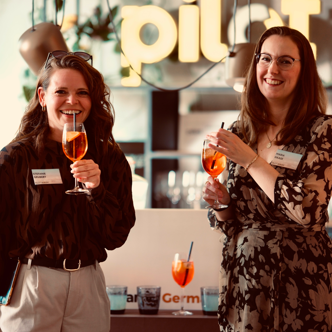 Zwei Frauen stehen lächelnd zusammen, halten Aperitifgläser in der Hand und genießen die Veranstaltung vor einem „pilot“-Leuchtschild im Hintergrund.