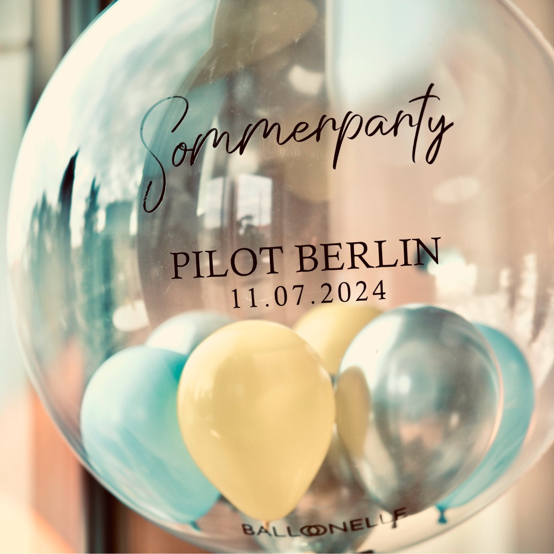 Ein transparenter Ballon mit der Aufschrift „Sommerparty PILOT BERLIN 11.07.2024“, gefüllt mit gelben und blauen Luftballons im Inneren.