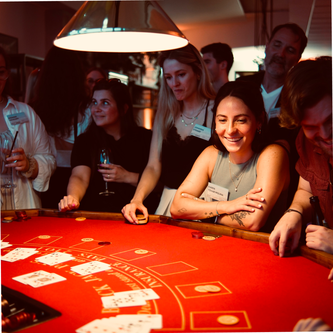 Eine Gruppe von Menschen steht um einen Roulette-Tisch. Eine Frau lächelt, während sie zuschaut, und andere halten Getränke in der Hand.