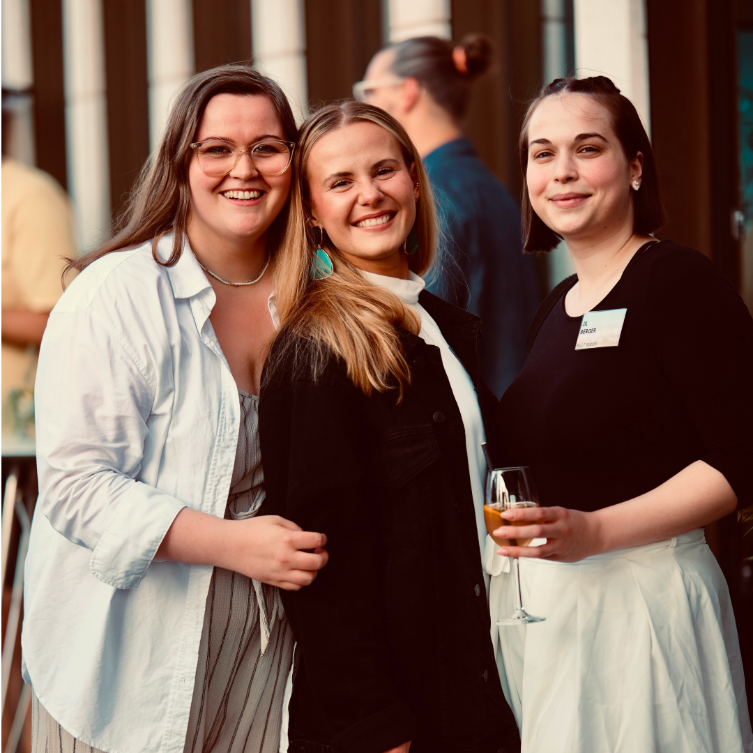 Drei Frauen stehen eng beieinander und lächeln in die Kamera, während eine von ihnen ein Glas in der Hand hält.